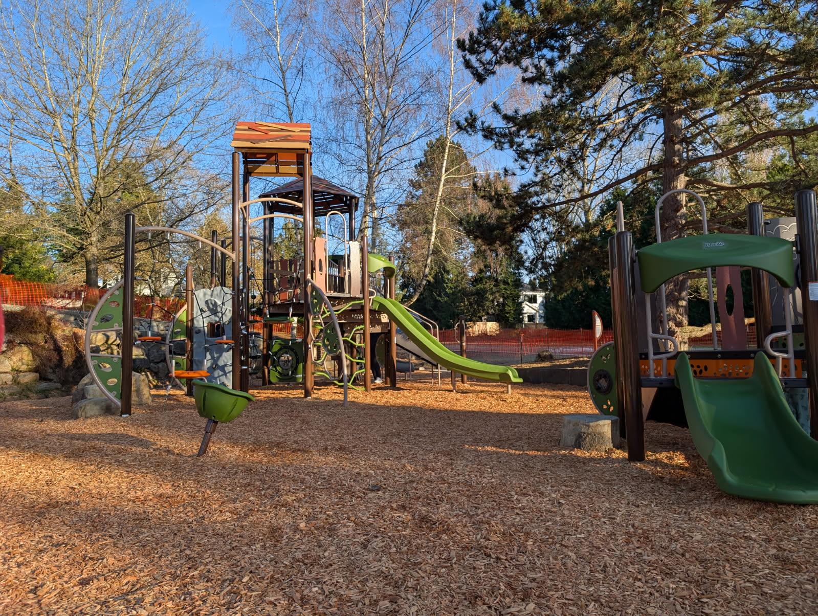 A wide view of a multi-component playground with green and brown equipment, including slides, climbing ropes, and a tall tower. The structure sits on a bed of wood chips, surrounded by tall deciduous and evergreen trees. Orange safety fencing is visible in the background against a bright, sunny sky.