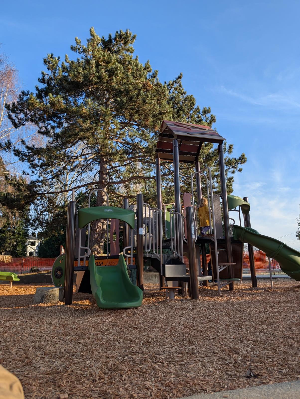 A modern playground structure featuring a small green slide in the foreground and a taller climbing tower with a brown roof in the background. A child in a yellow coat is visible playing on the upper level. The play area is covered in wood chips and framed by a large, mature evergreen tree under a clear blue sky.