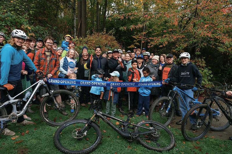 A large group of community members and cyclists gather with mountain bikes for the Cheasty trail ribbon-cutting event in 2025, standing outdoors in a wooded park and holding a ‘Grand Opening’ ribbon.