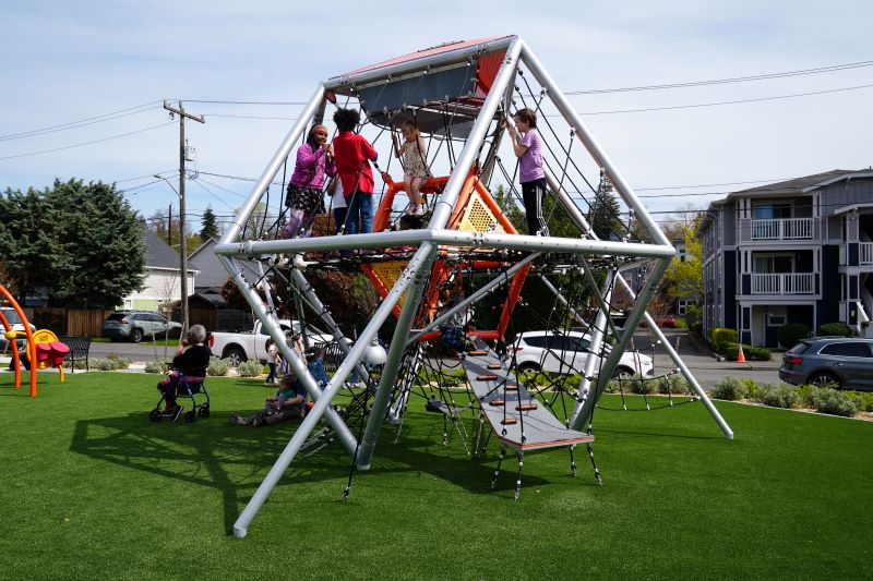 A geometric climbing structure made of metal and ropes rises above synthetic turf as children climb and explore it. Adults and children gather nearby, and residential buildings and trees line the park perimeter.