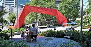 A man sits at a small table outdoors in a park dominated by a large red sculpture.