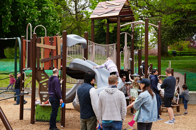 Children and adults enjoy various climbing and sliding elements on an expansive playground, with nature-themed panels