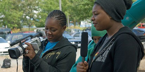 A teenager filming a subject while her friend uses a microphone