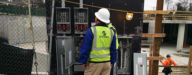 A construction worker wearing safety gear inspects open electrical panels at an active construction site, with fencing and partially built structures in the background.