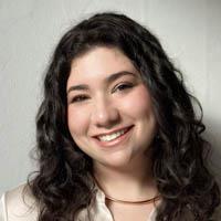 Sara smiling and has dark brown, curly hair, wearing cream color button up shirt.