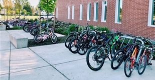 A row of children's bikes are locked to a long bike rack that runs the length of a brick wall outside an elementary school.