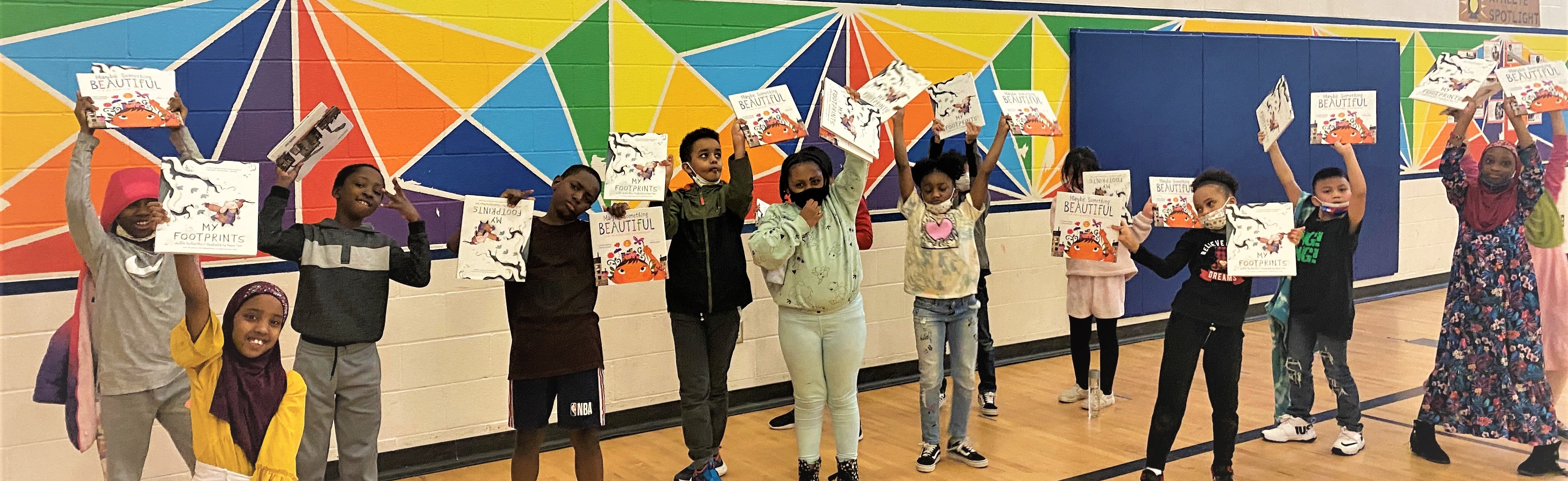 A group of elementary age children stand together in a school gym and hold up picture books while smiling.