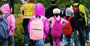 A group of children wearing brightly colored coats and backpacks walk down a sidewalk on their way to school with adults wearing Vision Zero safety vests.