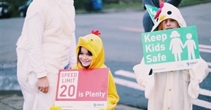 Two school age children wearing chicken onesies hold Vision Zero signs that say "20 is plenty" and "Keep Kids Safe."