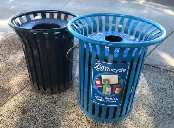 Photo of two fluted style cans with a wider upper rim and vertical side slats, a blue recycling can in foreground on thr right, and a black garbage can in back on left.