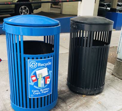 Photo of two of the most common public litter cans with rounded tops and vertical metal slats on sides, a blue recycling can on left and black garbage can on right.