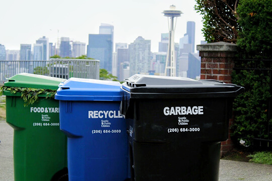 Photo of Seattle's residential waste containers including compost (green), recycling (blue), and garbage (black) in front of Seattle skyline.