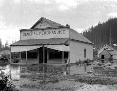 The General Merchandise store and other wooden buildings of Moncton are surrounded by large water puddles and wet ground.