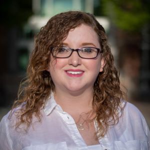 Headshot for SDC commissioner Taylor Woods. Taylor has curly shoulder length hair and is wearing black glasses. Her shirt is a white button up. The background is a blurred treescape.