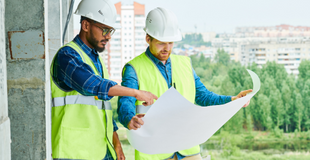 Two men wearing bright safety vests and white hard hats hold out a blueprint in front of a construction zone.