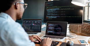 Man wearing glasses sits at a desk and types code onto his laptop while a larger monitor displays the code.