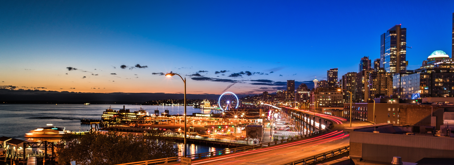 Evening view of a waterfront cityscape with illuminated buildings, a Ferris wheel, highways with light trails, and a calm body of water at sunset.