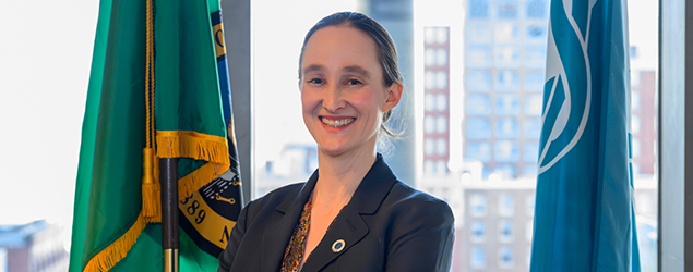 Mayor Katie B. Wilson smiling, in a dark blazer stands with her arms crossed in an office setting, with two flags behind her and a city skyline visible through the windows
