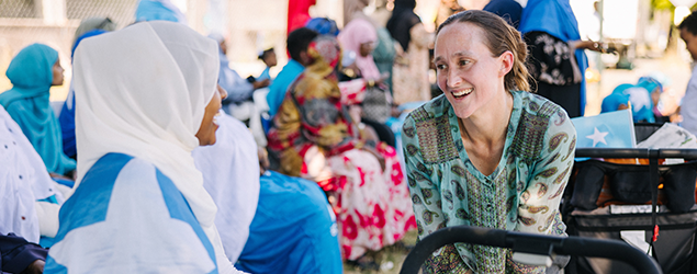 Mayor Katie B. Wilson leans toward and talks with a woman wearing a white headscarf, while other women in colorful clothing sit and stand together in the background at an outdoor gathering.
