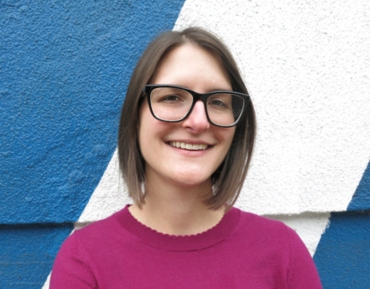 McCaela smiling at the camera in glasses and a magenta shirt in front of a bright blue and white painted wall.